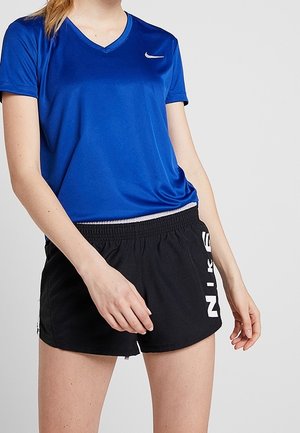 Person trägt ein blaues Nike-T-Shirt mit kurzen Ärmeln und schwarze Nike-Shorts mit weißem Logo an der Seite, steht mit entspannten Händen.