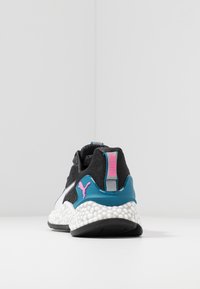 Sneaker Puma noir et blanc avec un soutien de talon bleu et un logo rose, semelle blanche texturée, vue de l'arrière sur fond blanc.