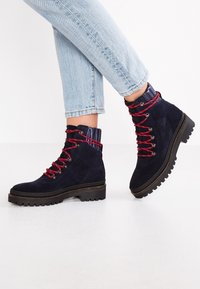 Bottes basses en daim bleu marine avec lacets rouges et argentés, dotées d'un talon texturé et d'une semelle extérieure noire épaisse.