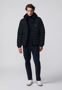 Giacca a vento nera con cappuccio, design trapuntato, zip frontale e patch del logo. Indossata sopra un dolcevita blu navy e abbinata a pantaloni scuri e sneakers.