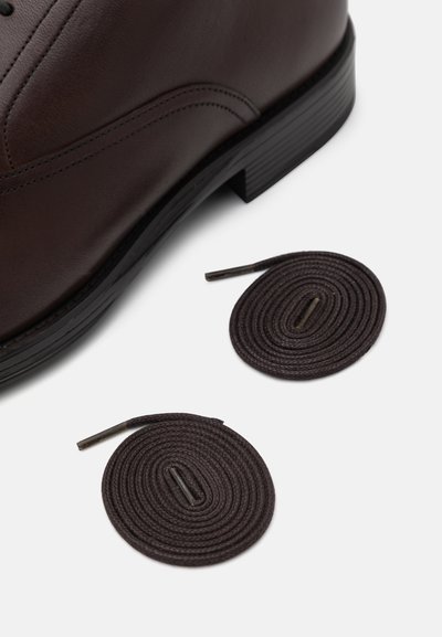 Scarpa in pelle marrone con punta arrotondata e superficie testurizzata, accompagnata da due lacci marroni scuri avvolti in cerchio.