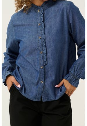 Kobieta ubrana w niebieską dżinsową koszulę na guziki z falbankami i elastycznymi mankietami, w połączeniu z czarnymi spodniami, z rękami w kieszeniach.