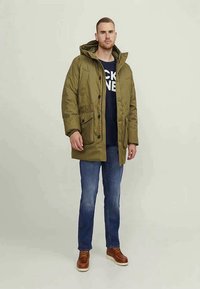 Casaco acolchoado verde-oliva com capuz, botões frontais e dois bolsos grandes, combinado com uma t-shirt azul-marinho, jeans azuis e sapatos castanhos.