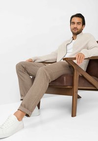 Lys beige ribbet cardigan over en hvid t-shirt, parret med brune bukser og hvide sneakers, siddende i en træstol.