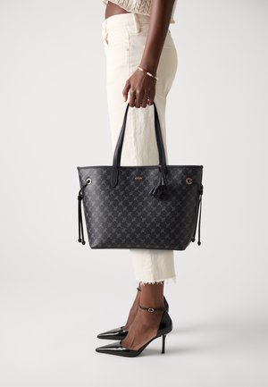 A person stands holding a stylish black tote bag with a patterned design, wearing beige pants and black high heels.