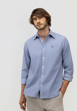 Hombre con cabello castaño que lleva una camisa azul claro abotonada con las mangas remangadas y pantalones beige, de pie frente a un fondo liso.