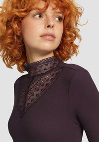 Dunkelviolette Bluse mit hohem Spitzkragen und geometrischen Mustern. Der Stoff hat eine gerippte Textur, die eine figurbetonte Silhouette betont.