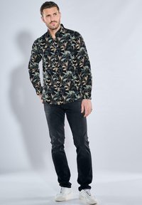 Schwarzes Langarmhemd mit Knopfleiste, das ein farbenfrohes Blumenmuster zeigt, kombiniert mit dunklen Jeans und weißen Sneakern. Glatte Textur, modern geschnitten.