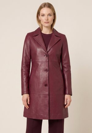 Kvinde iført en knaplukket, knælængde bordeaux skindfrakke over matchende bukser, stående mod en ensfarvet beige baggrund.