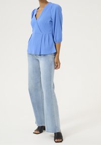 Femme portant un chemisier cache-cœur bleu clair à manches trois-quarts, un jean large délavé clair et des chaussures à talons noires à bout ouvert.