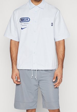 Man wearing a white short-sleeve button-up shirt with NBA and Nike logos and gray knee-length shorts.