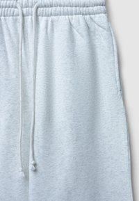 Un pantalon de survêtement gris clair avec une texture douce, une taille élastique et un cordon de serrage réglable. Dispose de poches latérales des deux côtés.