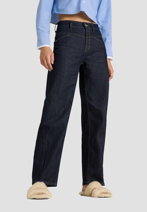Person trägt dunkelblaue Jeans mit geradem Bein, hellblaues kurz geschnittenes Hemd und beige flauschige Hausschuhe und steht vor einem einfarbigen Hintergrund.
