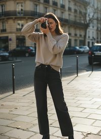 Helles beiges Sweatshirt mit Perlendetails an den Ärmeln, kombiniert mit hochgeschnittenen dunklen Jeans. Das Model hält eine Kamera, draußen, mit Bürgersteig und Gebäuden im Hintergrund.