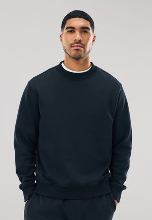 Fiatal férfi rövid fekete hajjal és ápolt szakállal egyszerű fekete pulóvert és fekete nadrágot visel, világosszürke háttér előtt áll.