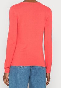 Pull coral à manches longues avec une texture côtelée et un col rond, porté sur un jean en denim bleu clair, mettant en avant une coupe décontractée.