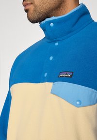 Φλις μπλούζα με κοντό φερμουάρ, σε μπλε και εκρού χρώμα, με ψηλό γιακά, κουμπώματα και τσέπη στο στήθος. Διαθέτει λογότυπο Patagonia. Μαλακή υφή.