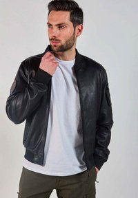 Veste bomber en cuir noir avec des poignets et un ourlet côtelés, dotée d'une fermeture éclair à l'avant et d'une poche sur la manche. Portée sur un t-shirt blanc.