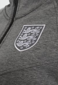 Sudadera gris con cremallera que presenta un emblema prominente con tres leones en un diseño de escudo en el pecho, tejido texturizado y corte deportivo.