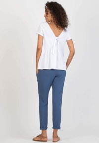 White peplum top with a deep V-back and tie detail, paired with loose-fitting blue pants and flat sandals. Smooth fabric texture.