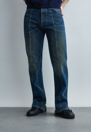 Άτομο που φοράει μπλε σκούρο τζιν με ίσια γραμμή, εμφανείς ραφές και ξεφτισμένα τελειώματα, συνδυασμένο με μαύρα casual παπούτσια σε γκρι δάπεδο.