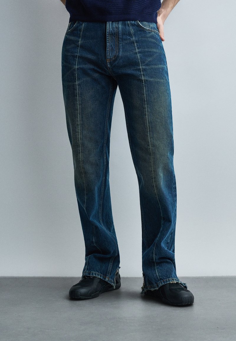 Persoană purtând blugi drepti de culoare albastru închis, cu cusături vizibile și tivuri destrămate, asortați cu pantofi casual negri pe podea gri.