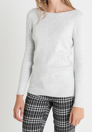 Kvinde iført en lysegrå ribstrikket langærmet trøje og sort-hvid ternede bukser, stående foran en ensfarvet baggrund.