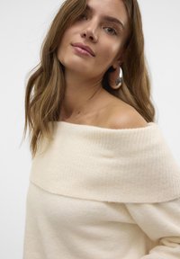 Crème off-the-shoulder trui van zacht breimateriaal, met een geribbelde textuur en een losse pasvorm. Zilveren ringoorbellen als opvallende accessoire.