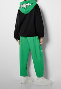 Schwarzer, übergroßer Kapuzenpullover mit grünem Kapuzenfutter und Grafik; grüne Jogginghose; weiße Sneakers. Entspannte Passform; weicher Stoff; sichtbare Nähdetails.