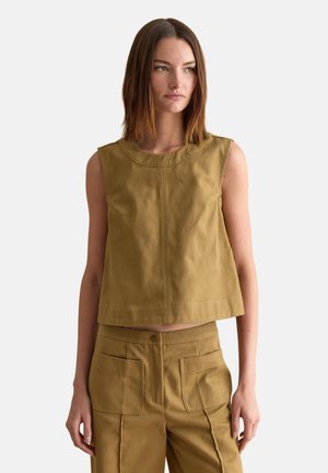 Femme aux cheveux bruns raides portant un haut court sans manches couleur beige et un pantalon taille haute assorti avec de grandes poches frontales, debout devant un fond blanc.