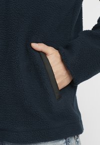 Navy blue fleece pullover with a textured surface, featuring a side pocket trimmed in black fabric. Hand partially visible in the pocket.
