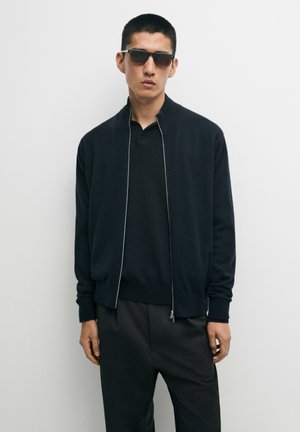 Man draag een zwarte jas met rits, zwart poloshirt, zwarte broek en donkere zonnebril, staand tegen een effen witte achtergrond.