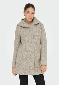 Manteau beige texturé avec une grande capuche, des poches avant et une silhouette légèrement ajustée. Tissu lisse avec un motif moucheté tout au long.