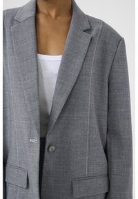 Harmaa bleiseri, jossa on teksturoitu kangas, lovetut käänteet, kaksi etutaskua ja valkoiset kontrastitikkaukset, yksittäisellä nappikiinnityksellä.