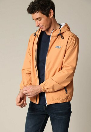 Lichtoranje regenjack met capuchon, rits voorkant en elastische manchetten. Heeft een klein logo-opdruk op de borst en een witte voering.