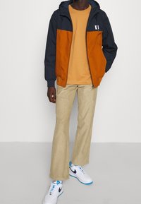 Vindjacka med marinblå och orange paneler, beiga manchesterbyxor och vita sneakers med blå accenter, bärs över en ljus senapsgul t-shirt.