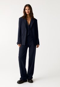 Marineblå stribet jakkesæt med en lang blazer og en dobbeltrækket vest. Bukser med bred benvidde. Glat tekstur og skræddersyet pasform. Sorte kraftige sko.