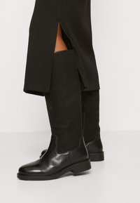 Schwarze kniehohe Stiefel mit einer glatten Lederzehe und einem Schaft aus Leinen. Mit einem seitlichen Reißverschluss und einer niedrigen, flachen Gummisohle. Strukturierte Oberfläche.