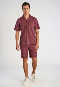 Camicetta bordeaux a maniche corte e pantaloni corti abbinati con un motivo testurizzato. Il tessuto ha una leggera finitura stropicciata. Indossata con sneakers bianche.