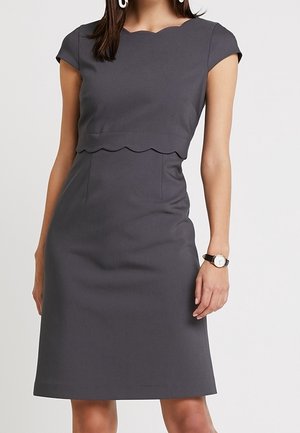 Femme portant une robe gris foncé ajustée à manches courtes avec des bords festonnés au niveau du décolleté et de la taille, accessoirisée d'une montre-bracelet.