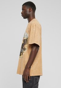 Oversized beige grafisk t-shirt lavet af blødt bomuld, med et ørnedesign og en falmet tekstur. Vist fra siden og med korte ærmer.