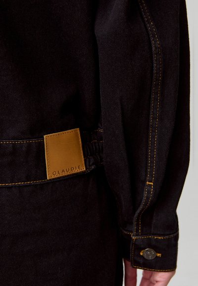 Primo piano della manica e della vita di una giacca di jeans nera con una toppa in pelle marrone etichettata "Claudie" e cuciture gialle a contrasto.