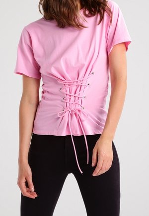 Femme portant une chemise rose à manches courtes avec un détail de laçage style corset sur le devant et un pantalon noir sur un fond uni.