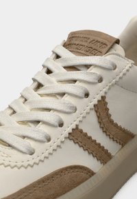 Αθλητικά παπούτσια με κρεμ δερμάτινο πάνω μέρος, λεπτομέρειες από σουέτ σε καφέ απόχρωση, υφή ραφής και λευκά κορδόνια. Διαθέτουν επίσης μάρκα με καφέ ταμπέλα στη γλώσσα.