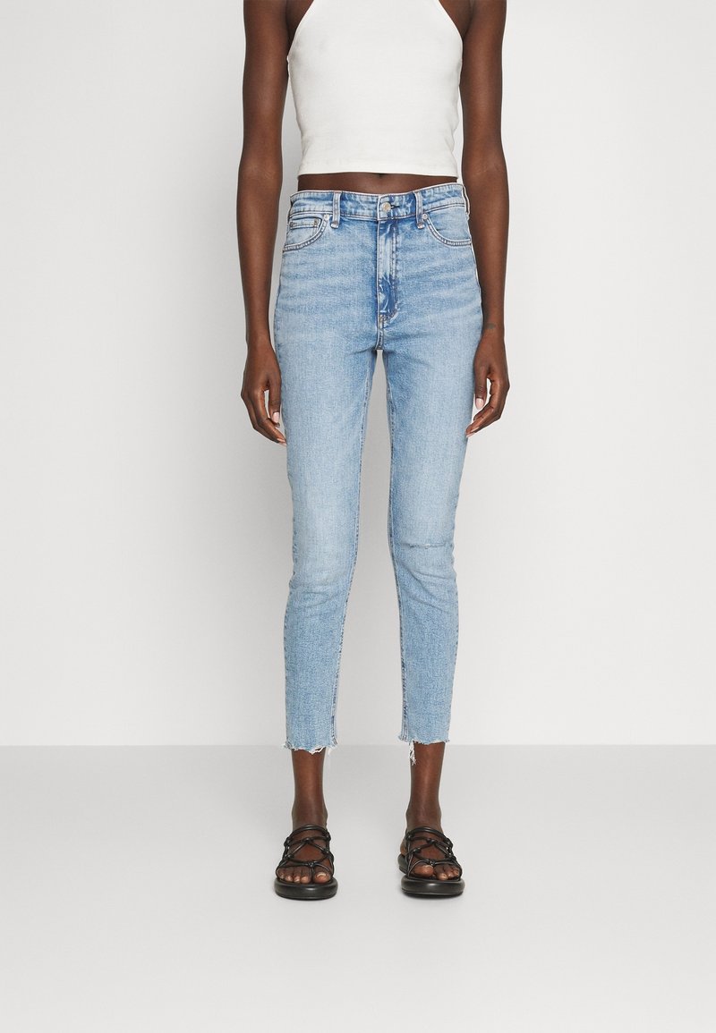 Jean bleu clair taille haute, coupe slim avec ourlet effiloché. Associé à un top court blanc à col halter et des sandales noires à brides.