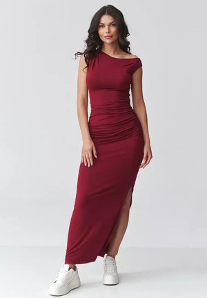 Kobieta z długimi ciemnymi włosami nosi dopasowaną bordową sukienkę z rozcięciem z boku oraz białe platformowe trampki, stojąc na tle jasnego, jednolitego tła.