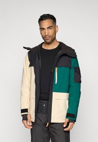 Casaco isolante com um design multicolorido em creme, preto, verde e azul marinho, apresentando capuz, vários bolsos e punhos ajustáveis.