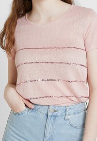 Różowy t-shirt z krótkim rękawem wykonany z miękkiej, teksturowanej tkaniny z poziomymi pasami z cekinów. W zestawie z jasnoniebieskimi, wysokim stanem jeansami.