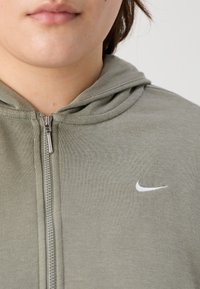 Sudadera con capucha gris con textura suave, capucha y un logo blanco de Nike en el lado izquierdo del pecho. La cremallera y las costuras son visibles.