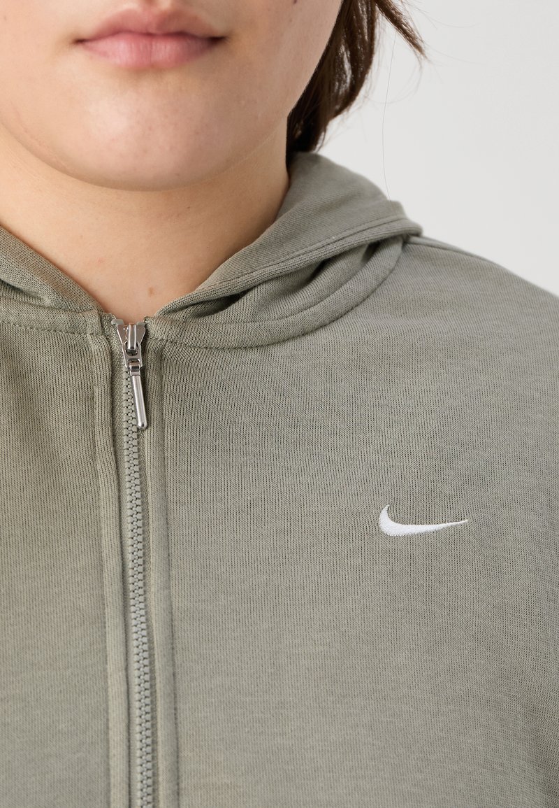 Sudadera con capucha gris con textura suave, capucha y un logo blanco de Nike en el lado izquierdo del pecho. La cremallera y las costuras son visibles.